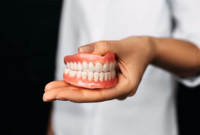 Hand holding dental model showing full set of artificial teeth against black background.