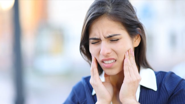 Woman experiencing tooth pain, touching her cheek with both hands in discomfort outdoors.